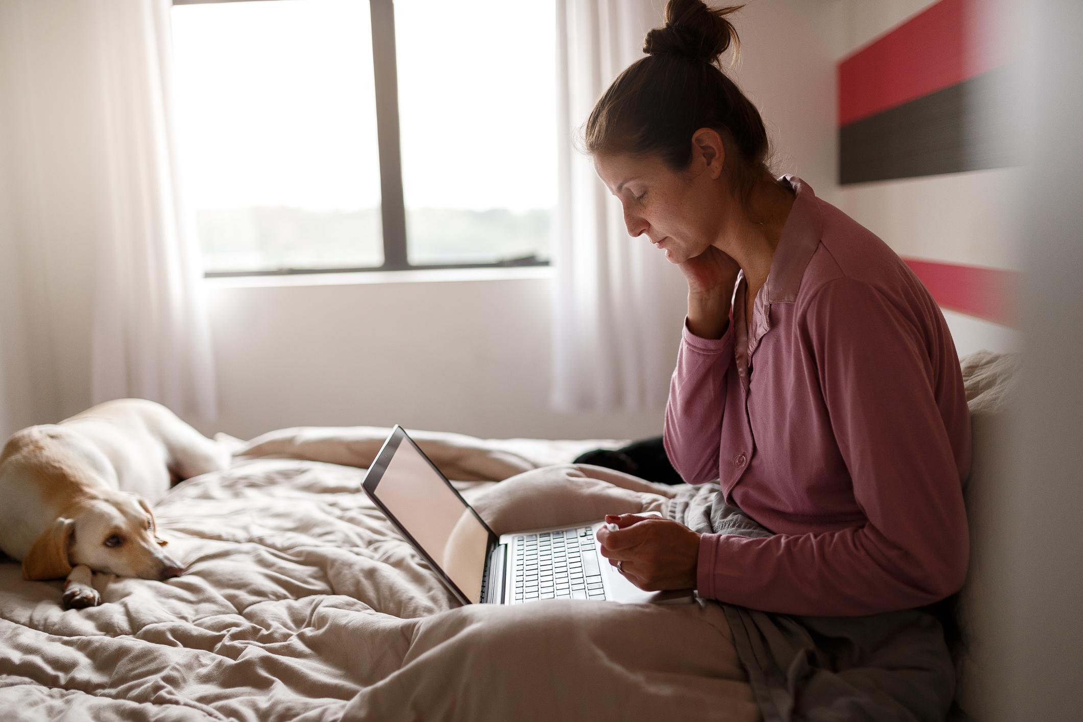 Eine Frau sitzt auf einem Bett in einem hellen Schlafzimmer und schaut auf ihren Computer hinunter. Ein Hund liegt neben ihr auf dem Bett.